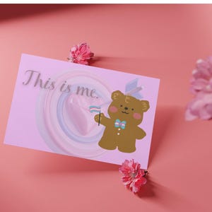May include: A pink greeting card with the text "This is me" and a cartoon bear holding a flag with the transgender pride colors. The card is decorated with pastel swirls and placed on a pink surface, with pink flowers scattered around.
