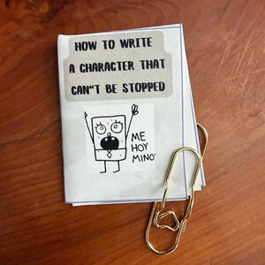 May include: A small booklet with the text "HOW TO WRITE A CHARACTER THAT CAN'T BE STOPPED" and a cartoon drawing of a character. A gold-coloured paper clip holds the booklet together. The booklet is on a wooden surface.