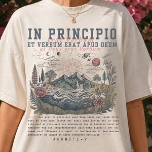 May include: An ivory short-sleeve t-shirt featuring a graphic print with the text IN PRINCIPIO ET VERBUM ERAT APUD DEUM and JOHN 1:1-7. The illustration depicts a mountain landscape with a sun, moon, stars, trees, flowers, and a waterfall.