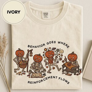 Könnte beinhalten: Ein elfenbeinfarbenes T-Shirt mit einer Grafik von vier Skeletten mit Kürbisköpfen. Der Text lautet "BEHAVIOR GOES WHERE REINFORCEMENT FLOWS". Die Skelette sind dargestellt, wie sie einen Korb mit Süßigkeiten halten, lesen, tanzen und zeichnen.