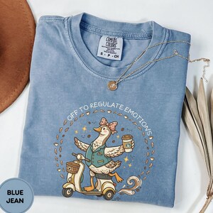 May include: A blue jean coloured t-shirt featuring a graphic of a goose wearing a vest and bow, riding a scooter while holding a coffee cup. The text reads OFF TO REGULATE EMOTIONS, surrounded by coffee beans and leaves. A thin gold chain necklace rests on the shirt.
