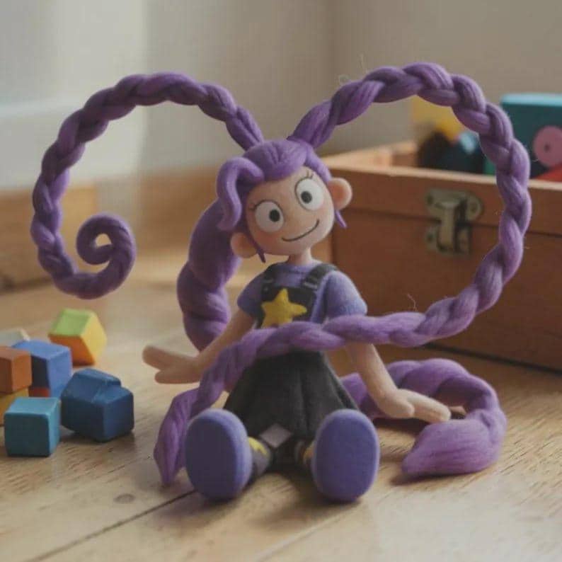 May include: A handcrafted doll with purple, looped, braided hair. The doll is dressed in a purple top with a yellow star, black dungarees, and a black skirt. It sits on a wooden floor, with colourful wooden blocks scattered around.