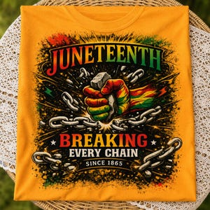 Op de afbeelding: Een mosterdgeel t-shirt met een afbeelding van een vuist die metalen kettingen breekt. De tekst luidt JUNETEENTH BREAKING EVERY CHAIN SINCE 1865. De vuist is gekleurd in rood, geel en groen en het ontwerp bevat zwarte verfspatten als effect.