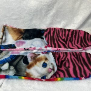 May include: A pair of cozy cat-themed slippers. The top slipper features a calico cat design, while the bottom slipper showcases a cat with blue eyes. The soles have a pink zebra print. The slippers are lined with a rainbow trim.