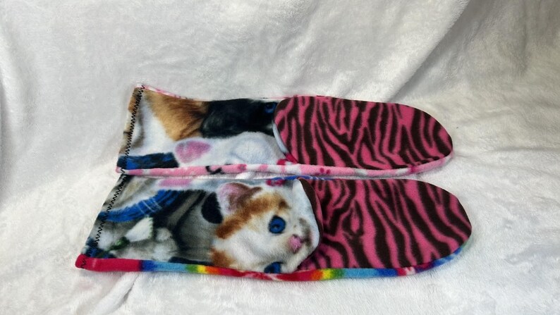 May include: A pair of cozy cat-themed slippers. The top slipper features a calico cat design, while the bottom slipper showcases a cat with blue eyes. The soles have a pink zebra print. The slippers are lined with a rainbow trim.