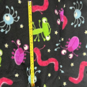 Puede incluir: Tela de forro polar negra con un patrón repetido de criaturas de dibujos animados coloridas y estrellas. Una cinta métrica amarilla muestra una longitud de aproximadamente 25 cm. Las criaturas son rosas, verdes, azules y rojas.