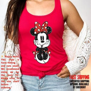 May include: Red tank top featuring a Minnie Mouse graphic. The graphic shows Minnie Mouse with a red bow with white polka dots. The tank top has a scoop neck and is form-fitting. The text on the image says, "Please Note: This style is form-fitting and runs small."