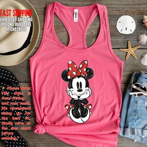 May include: A pink tank top featuring a Minnie Mouse graphic. The cartoon character is depicted with a red bow with white polka dots. The tank top has a racerback design and is made of a soft material.