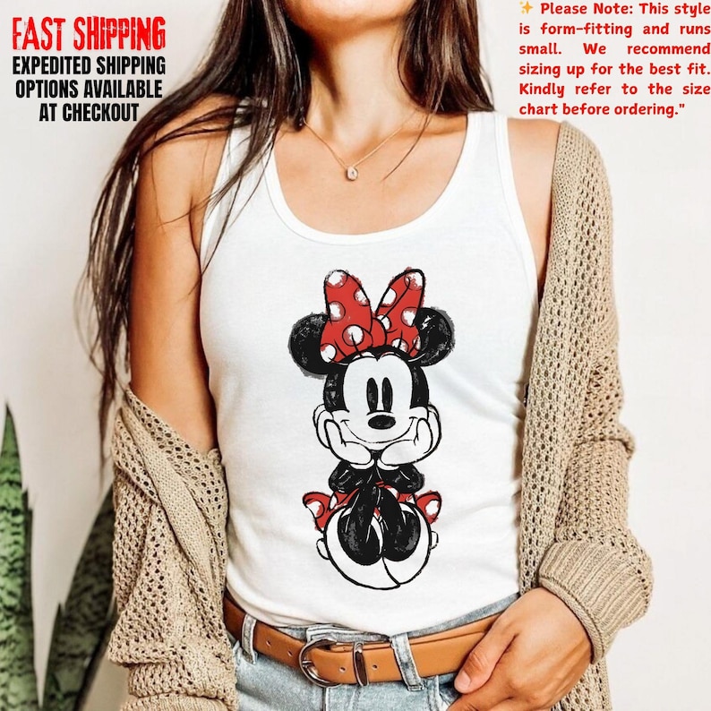 May include: White tank top featuring a Minnie Mouse graphic with a red polka dot bow. The image includes text that says "FAST SHIPPING" and "EXPEDITED SHIPPING OPTIONS AVAILABLE AT CHECKOUT". Additional text advises to refer to the size chart.