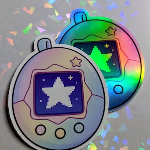 May include: Two holographic stickers in the shape of a retro handheld game. One sticker is a pastel purple and blue, the other is green and blue. Each has a star graphic on the screen and a star-shaped outline.