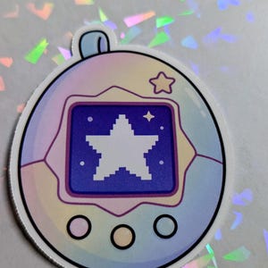 May include: A sticker featuring a retro-style digital pet device. The device has a blue screen displaying a white pixelated star. The casing is a gradient of pastel colors, with a small star icon at the top. Three black circles are at the bottom.