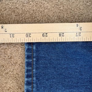 Op de afbeelding: Close-up van blauwe denimstof met een houten liniaal met afmetingen in inches. De liniaal geeft een meting van ongeveer 68,5 tot 78,7 cm aan. De stof heeft een zichtbare naad.
