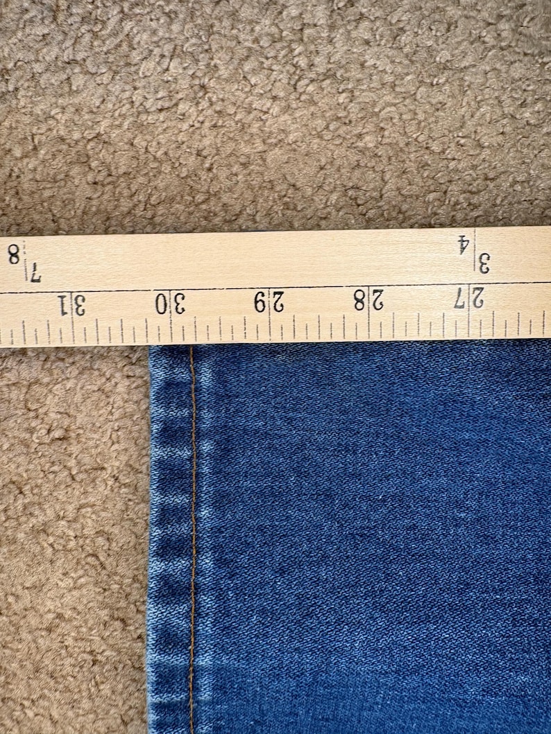 Op de afbeelding: Close-up van blauwe denimstof met een houten liniaal met afmetingen in inches. De liniaal geeft een meting van ongeveer 68,5 tot 78,7 cm aan. De stof heeft een zichtbare naad.