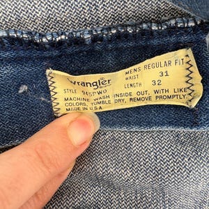 Op de afbeelding: Close-up van een Wrangler denim kledinglabel. Het gele label heeft zwarte tekst met een taillemaat van 31 inch (79 cm) en een lengte van 32 inch (81 cm). Wasvoorschriften en "Made in USA" zijn ook zichtbaar.