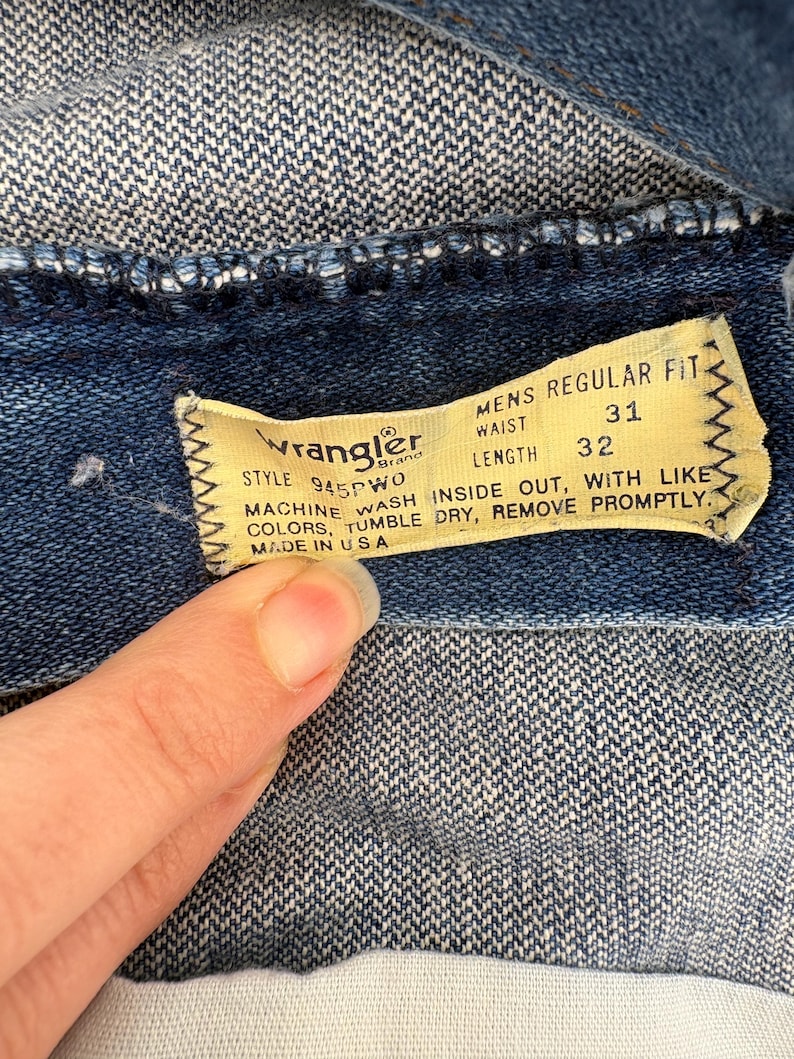 Op de afbeelding: Close-up van een Wrangler denim kledinglabel. Het gele label heeft zwarte tekst met een taillemaat van 31 inch (79 cm) en een lengte van 32 inch (81 cm). Wasvoorschriften en "Made in USA" zijn ook zichtbaar.