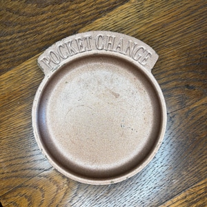 May include: A round, brown ceramic dish with the words "POCKET CHANGE" arched across the top. The dish has a shallow, circular interior and a slightly raised rim. The surface has a textured appearance.