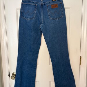 Op de afbeelding: Blauwe Wrangler jeans van denim met een klassieke pasvorm. De jeans heeft twee achterzakken, een bruine leren patch met het Wrangler-logo en een metalen ritssluiting. De jeans hangen aan een zwarte hanger.