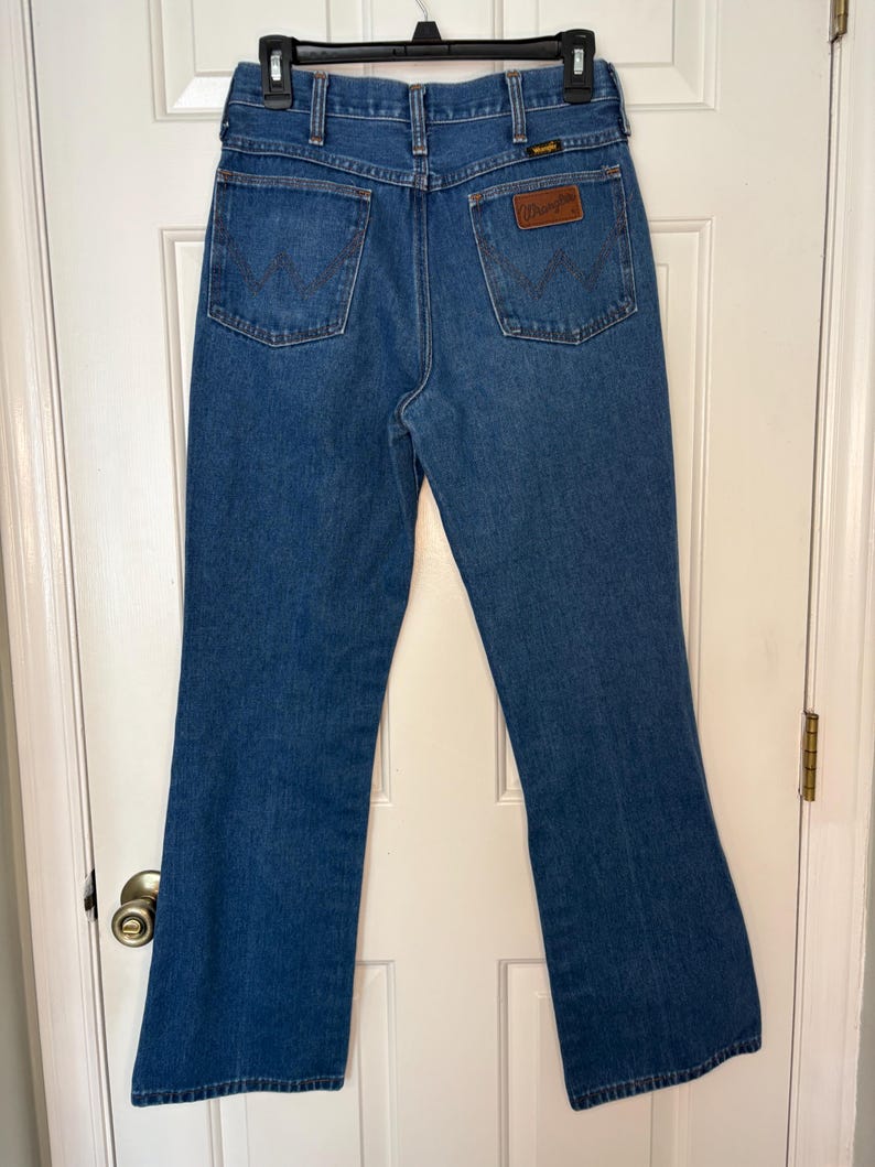 Op de afbeelding: Blauwe Wrangler jeans van denim met een klassieke pasvorm. De jeans heeft twee achterzakken, een bruine leren patch met het Wrangler-logo en een metalen ritssluiting. De jeans hangen aan een zwarte hanger.