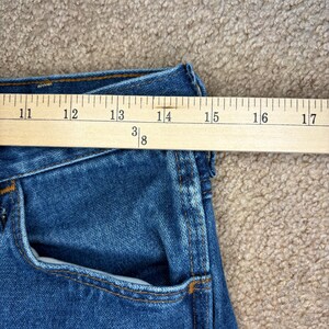 Op de afbeelding: Close-up van een blauwe spijkerbroek met een houten liniaal die de taille meet. De liniaal toont metingen van 28 tot 43 cm. De jeans heeft een klassiek ontwerp met vijf zakken en oranje stiksels.