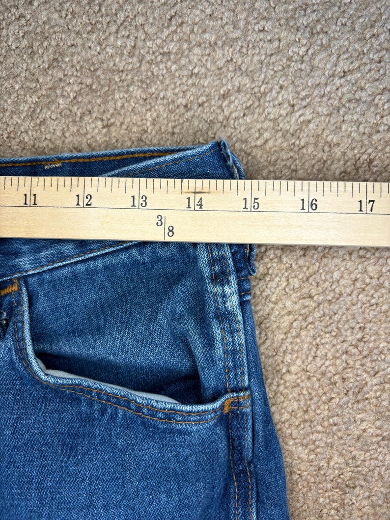 Op de afbeelding: Close-up van een blauwe spijkerbroek met een houten liniaal die de taille meet. De liniaal toont metingen van 28 tot 43 cm. De jeans heeft een klassiek ontwerp met vijf zakken en oranje stiksels.