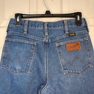 Op de afbeelding: Blauwe Wrangler jeans, achteraanzicht. Voorzien van het Wrangler-logo op een bruine leren patch en de kenmerkende W-vormige stiksels op de achterzakken. De jeans hangt aan een zwarte hanger.