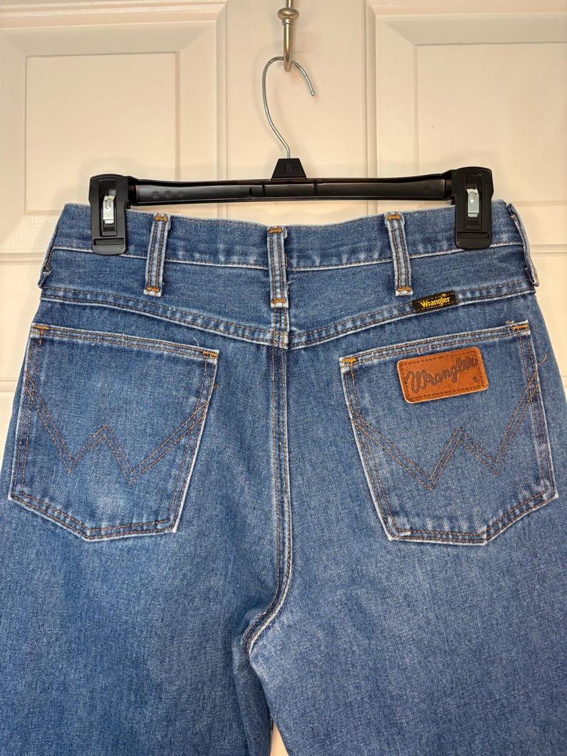 Op de afbeelding: Blauwe Wrangler jeans, achteraanzicht. Voorzien van het Wrangler-logo op een bruine leren patch en de kenmerkende W-vormige stiksels op de achterzakken. De jeans hangt aan een zwarte hanger.