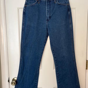 Op de afbeelding: Blauwe denim jeans met een klassiek ontwerp met vijf zakken en een lichte flare aan de zoom. De jeans hangen aan een zwarte hanger tegen een witte deur. De jeans zijn gemaakt van stevig denim materiaal.