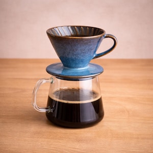 Puede incluir: Una cafetera de cerámica azul para verter con un platillo a juego se encuentra sobre una jarra de vidrio transparente llena de café oscuro. La cafetera tiene un asa y un interior texturizado. La configuración está sobre una superficie de madera.