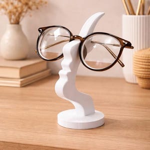 Puede incluir: Un soporte blanco para gafas con forma de rostro, sosteniendo un par de gafas con montura marrón. El soporte tiene una base redonda y un diseño curvo. Las gafas tienen lentes transparentes y patillas doradas. El soporte está sobre una superficie de madera.