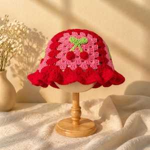 Può includere: Un cappello a secchiello all'uncinetto fatto a mano in rosso, rosa e verde. Il cappello presenta un design a ciliegia sulla corona e un bordo a balze. Il cappello è esposto su un supporto di legno. È un accessorio unico.
