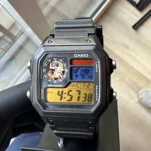 May include: A black Casio digital watch with a square case, featuring a world map display, a circular analog-style sub-dial with the text KIDS, and a yellow LCD screen showing the time 4:57:38. The watch is water resistant to 100 meters.