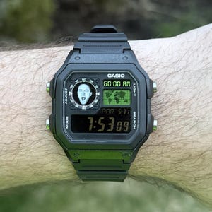 May include: Black Casio digital watch with a rectangular face. The watch displays the time, date, and a world map graphic. The watch face also features a small image of a person. The watch is worn on a wrist.