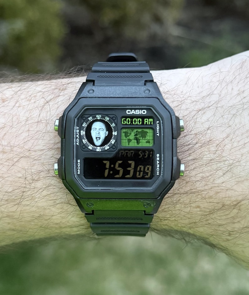 May include: Black Casio digital watch with a rectangular face. The watch displays the time, date, and a world map graphic. The watch face also features a small image of a person. The watch is worn on a wrist.