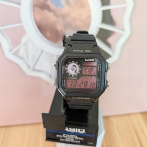 May include: Black Casio digital watch with a rectangular face, displaying the time and date. The watch has multiple buttons and a world time feature. The watch is on a black display stand.