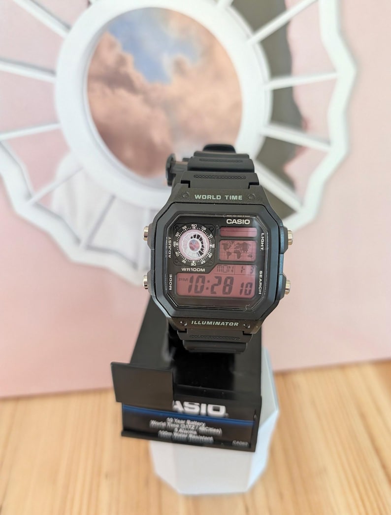 May include: Black Casio digital watch with a rectangular face, displaying the time and date. The watch has multiple buttons and a world time feature. The watch is on a black display stand.