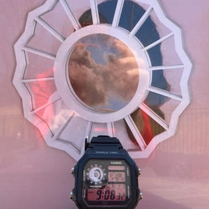 May include: Black Casio digital watch with a rectangular face, displaying the time 9:08:32. The watch is positioned in front of a white, scalloped-edged circular design with a cloudy sky reflected in the center.