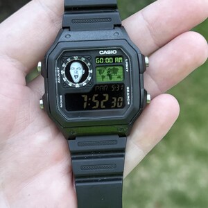May include: Black Casio digital watch with a black strap. The watch face shows the time, date, and a graphic. The watch is held in a hand.