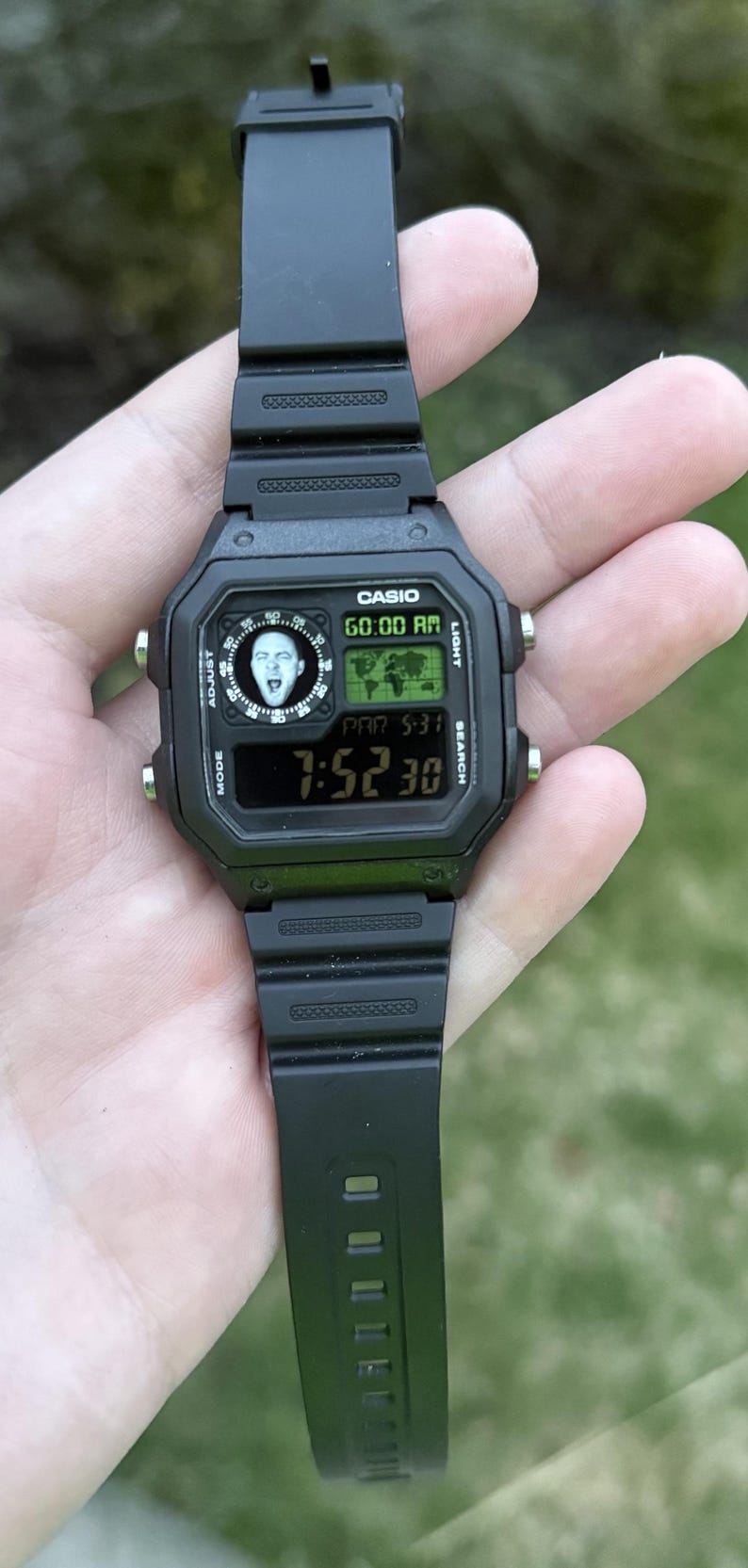 May include: Black Casio digital watch with a black strap. The watch face shows the time, date, and a graphic. The watch is held in a hand.