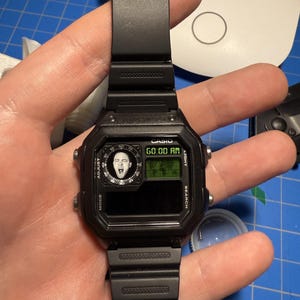 May include: Black Casio digital watch with a rectangular face and black strap. The watch face shows the time and a small image of a person's face. The watch has buttons for mode, adjust, search, and light.