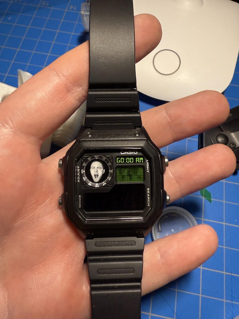 May include: Black Casio digital watch with a rectangular face and black strap. The watch face shows the time and a small image of a person's face. The watch has buttons for mode, adjust, search, and light.