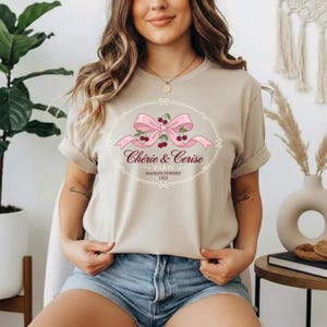 Op de afbeelding: Beige T-shirt met een roze strik en kersendesign en de tekst "Chérie & Cerise PARIS MAISON FONDÉE 1923". Het shirt heeft korte mouwen en een ronde hals.