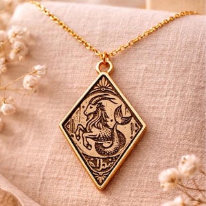 Puede incluir: Collar dorado con colgante en forma de diamante con el signo zodiacal de Capricornio grabado. El colgante representa una criatura mítica con cabeza de cabra y cola de pez. Cadena y colgante dorados.