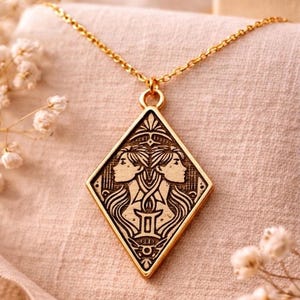 Puede incluir: Collar con colgante dorado con un dije en forma de diamante con un diseño del signo zodiacal Géminis. El dije muestra dos perfiles enfrentados, con detalles intrincados. El collar está sobre una tela suave de color beige rosado.
