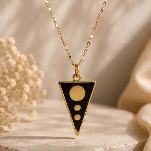 Op de afbeelding: Goudkleurige ketting met een driehoekige hanger. De hanger heeft een zwarte emaille achtergrond met een grote gouden cirkel en twee kleinere gouden cirkels. De ketting wordt gepresenteerd op een stenen oppervlak.