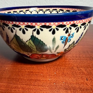 May include: A ceramic bowl with a floral and butterfly design. The bowl has a dark blue rim and a cream-colored interior. The exterior features a pattern of flowers, leaves, and a blue butterfly. The bowl is sitting on a wooden surface.