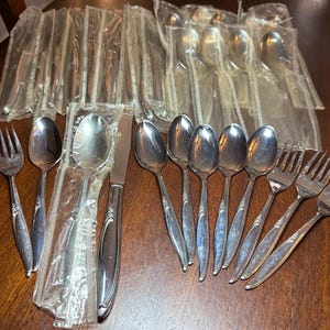 May include: A collection of stainless steel flatware including forks, spoons, and knives. Several pieces are individually wrapped in clear plastic sleeves, while others are unwrapped, displaying a polished silver finish and a floral handle design.