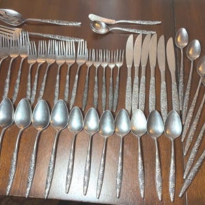 May include: A large collection of silver-toned flatware featuring a detailed floral and vine pattern on the handles. The set includes various forks, knives, spoons, a slotted serving spoon, and a butter knife, all arranged on a dark wooden surface.