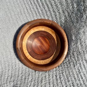 May include: A round wooden bowl with a multi-toned design. The bowl features concentric circles of varying wood shades, including dark brown, light brown, and a medium brown center. The bowl is likely for serving or display.