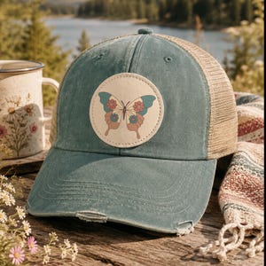 Op de afbeelding: Een verweerde blauwgroene honkbalpet met een beige mesh achterkant en een ronde patch op de voorkant. De patch bevat een grafische illustratie van een vlinder met blauwgroene, roze en gele vleugels versierd met kleine bloemmotieven op een crèmekleurige achtergrond.