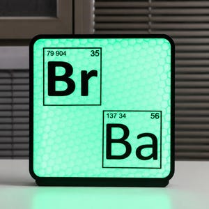 Op de afbeelding: Een vierkante lamp met een zwart frame en een groen oplichtend oppervlak. Het oppervlak toont de periodieke tabel symbolen "Br" en "Ba" in zwart, samen met hun atoomnummers en gewichten. De lamp staat op een wit oppervlak.